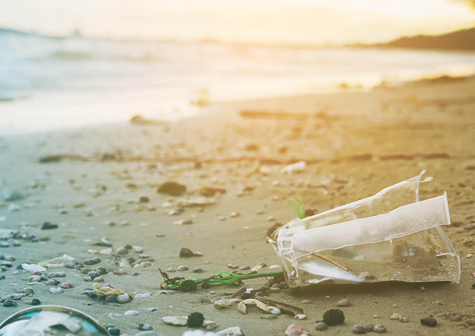 Beira mar ao por do sol com vários resíduos plásticos poluindo a paisagem, com destaque para uma garrafa plástica